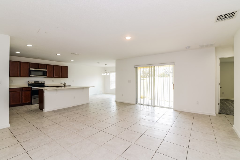 A spacious, empty living room with white tiled flooring and a kitchen area to the left.