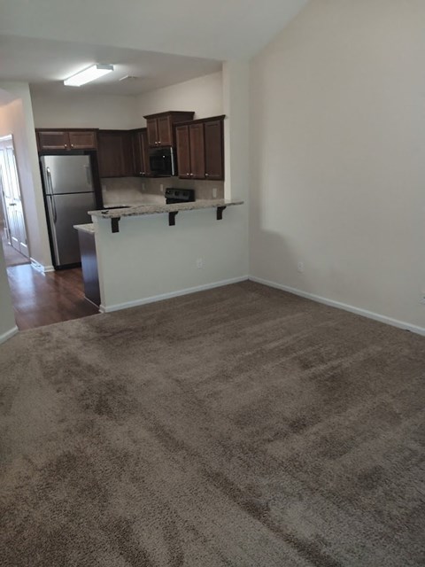 an empty living room with a kitchen and a carpeted floor