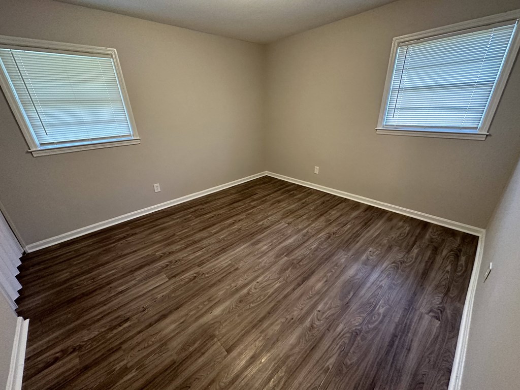A room with wooden flooring and two windows with blinds.