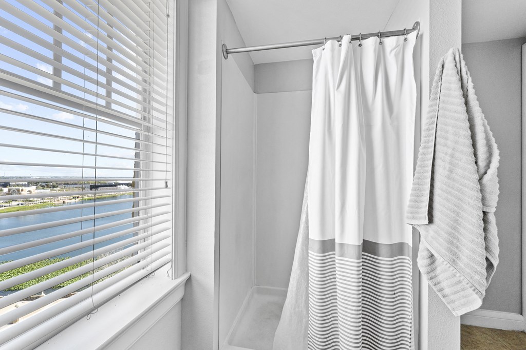 a bathroom with a shower and white curtains and a window with white blinds