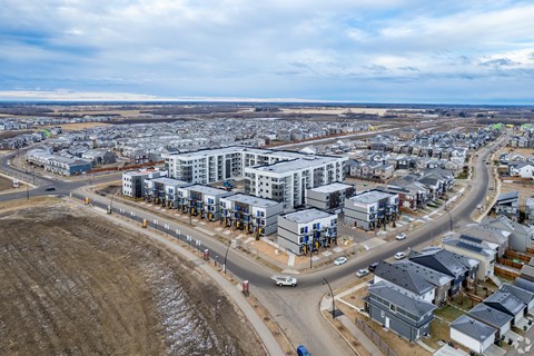 A large, modern residential complex is surrounded by a dirt field and other buildings.