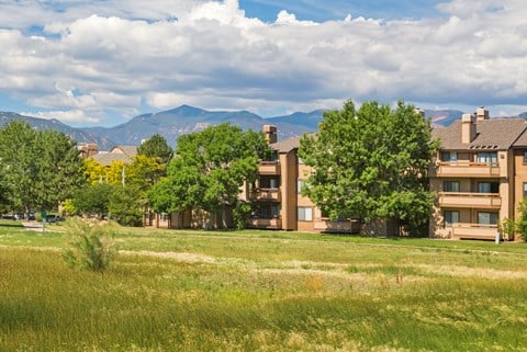 A grassy field in front of apartment buildings with trees and mountains in the background.