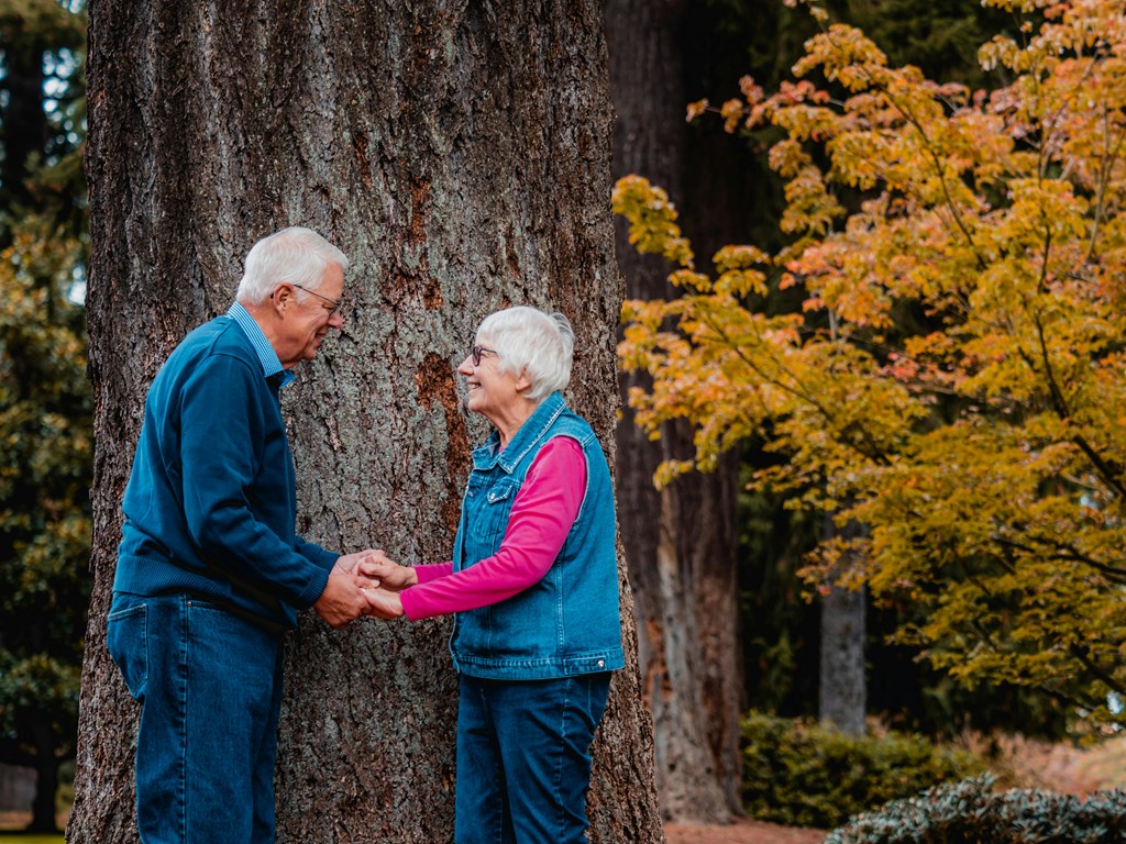 An elderly couple holding hands in front of a tree.