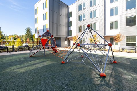 A playground with a red and grey climbing structure in front of a building.