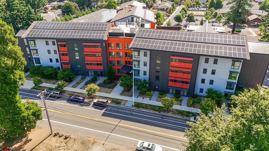 A modern building with solar panels on the roof is surrounded by trees and a street with cars.