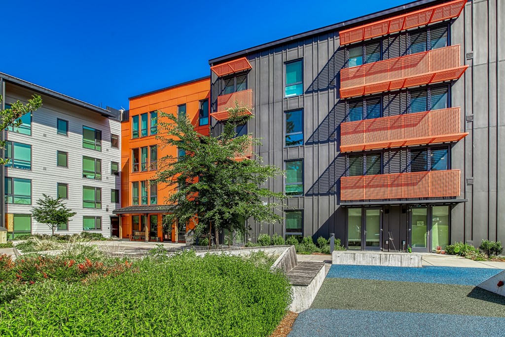 A modern building with orange balconies is surrounded by greenery and a small pond.