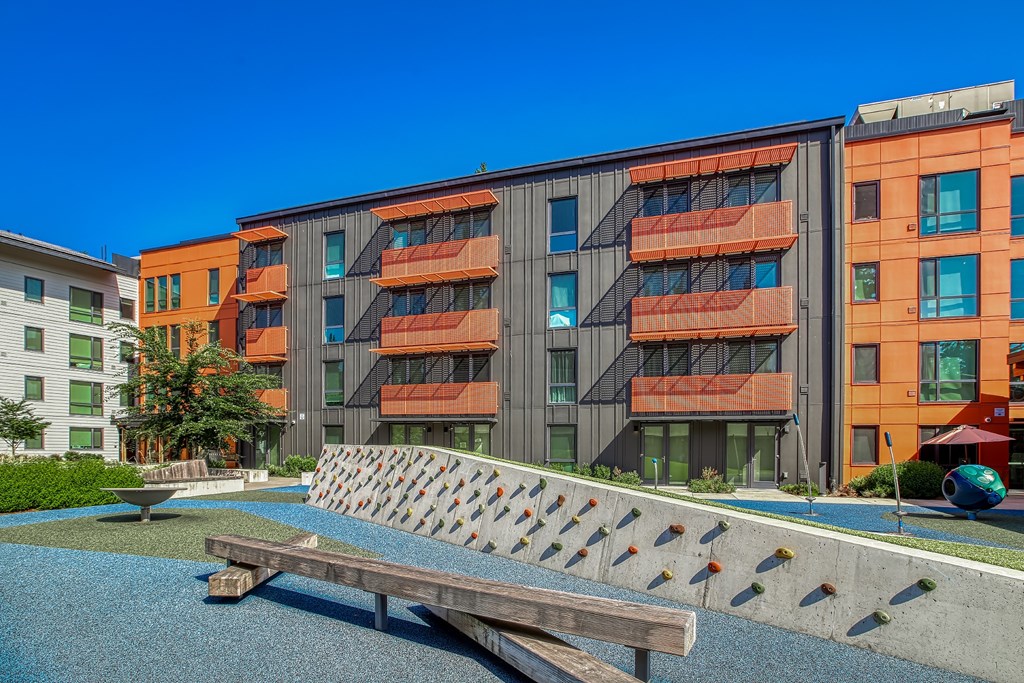A modern building with orange and grey balconies is surrounded by a small pool and a wooden bench.