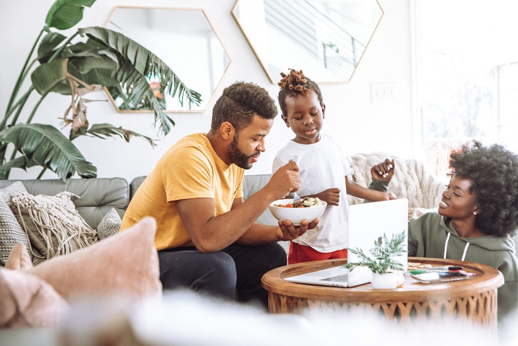 a man and woman sitting on a couch eating a bowl of food with a young girl standing