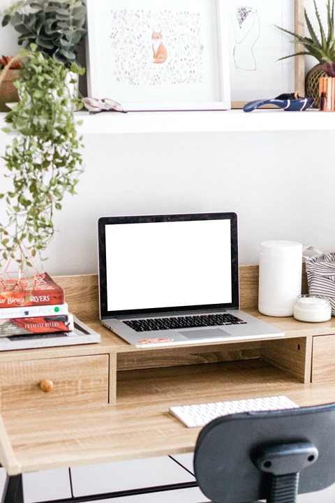 A laptop sits on a wooden desk with a white screen, surrounded by various objects including a potted plant, books, and a white vase.