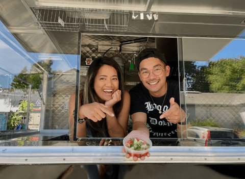 A man and woman are standing behind a food truck counter, smiling and pointing at the camera.