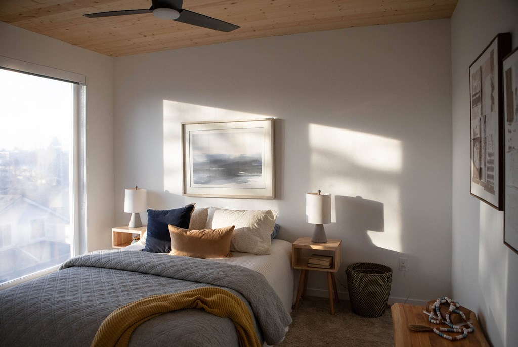 A bedroom with a bed, two lamps, a ceiling fan, and a painting on the wall.