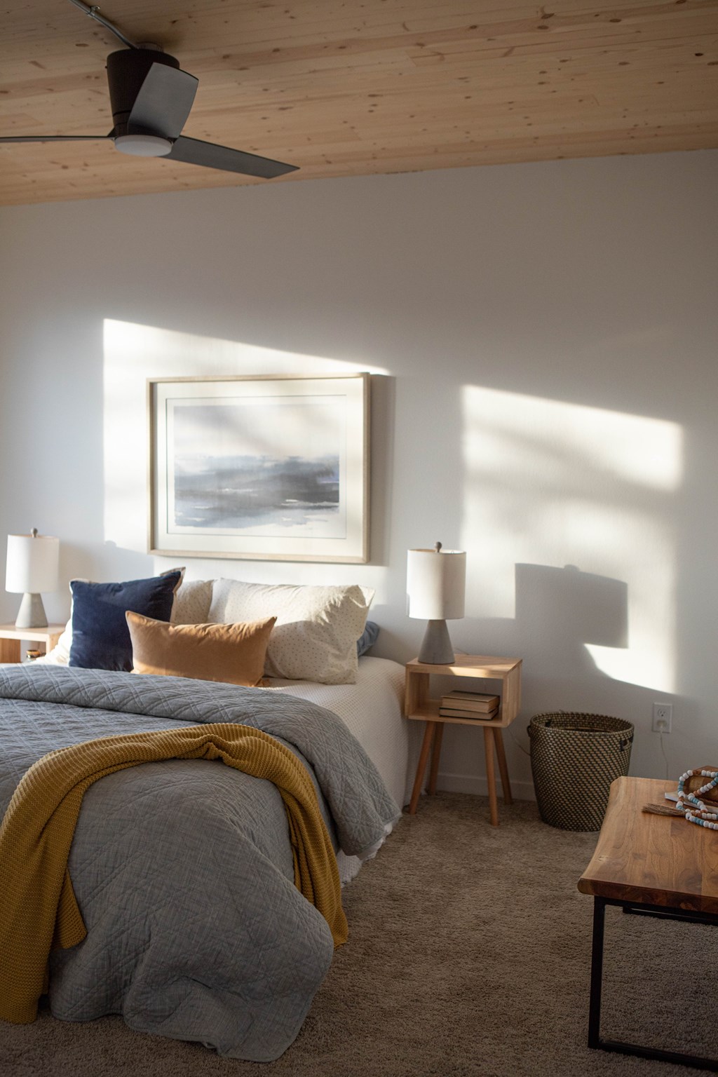 A bedroom with a bed, a nightstand, a lamp, a painting, a basket, and a table.