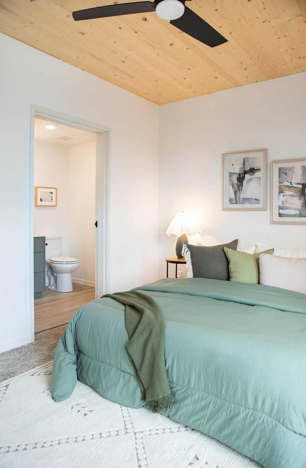 A bedroom with a bed, a fan, and a toilet in the background.