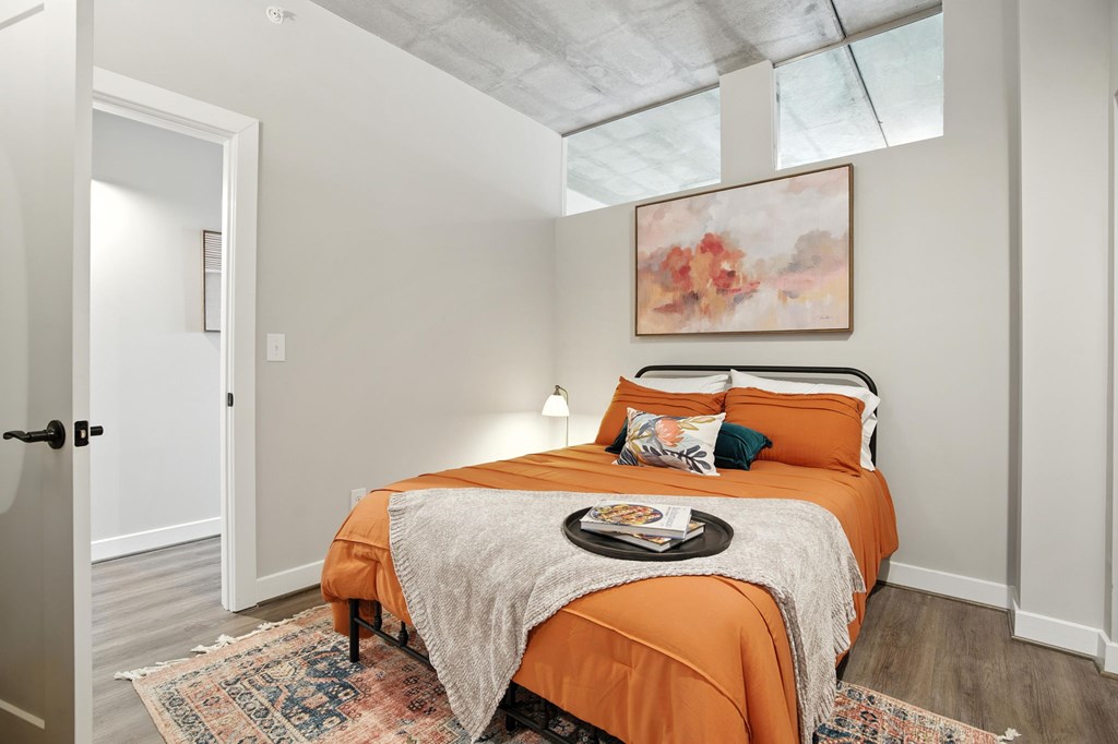 A bedroom with a bed covered in an orange bedspread and a painting above it.