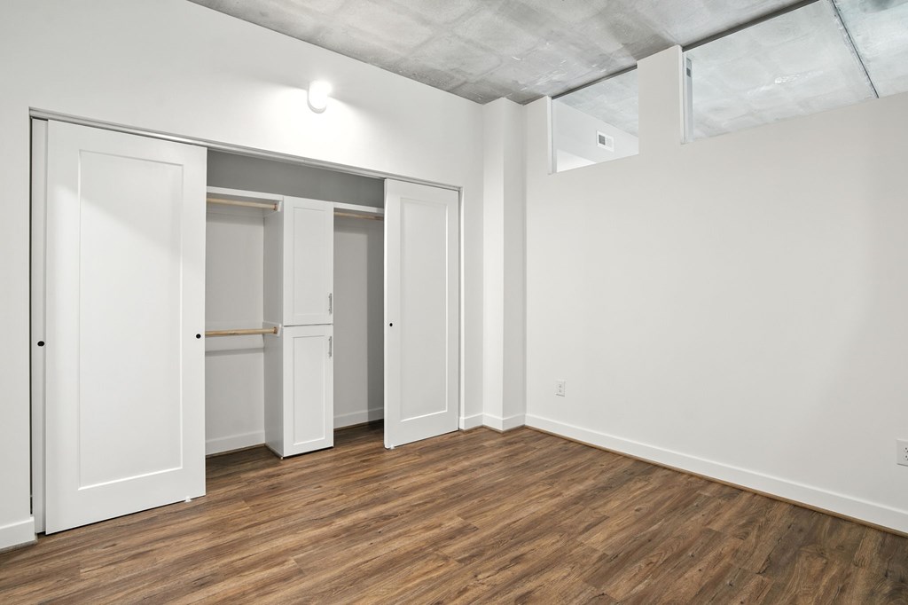 A room with white walls and wooden flooring, featuring a white door and a white closet with a bar across the door.
