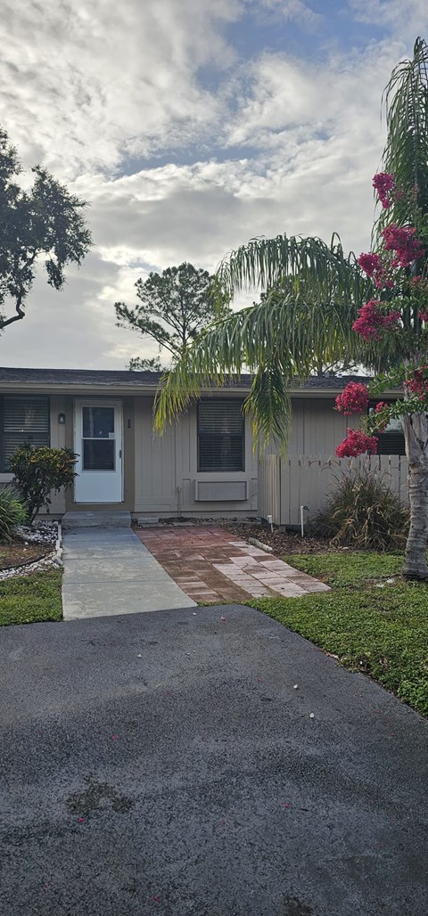 A house with a white door and windows is surrounded by greenery and a paved walkway.