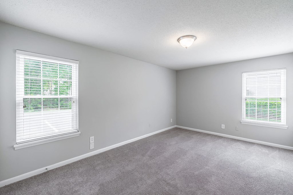 A room with a carpeted floor and two windows with blinds.