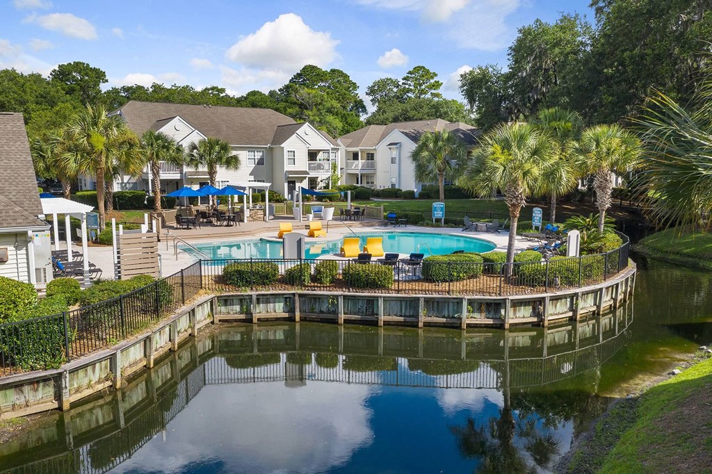 A pool surrounded by a fence and trees with houses in the background.