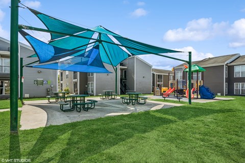 A playground with a blue and green umbrella and picnic tables.