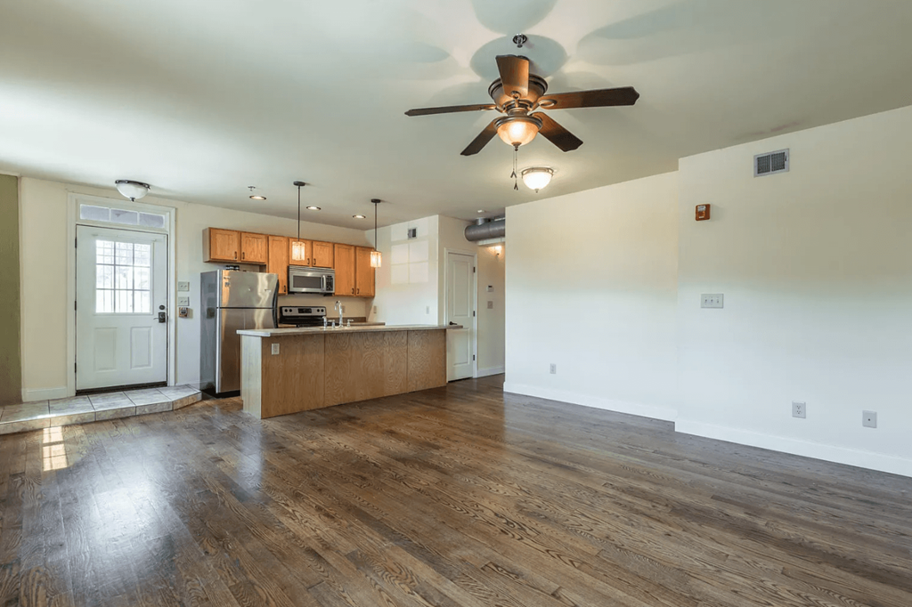 an empty living room with a ceiling fan and a kitchen in the background