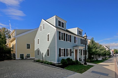 A white house with a yellow section on the side is in front of a row of trees.