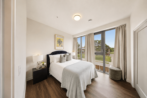 A bedroom with a large bed, a nightstand, curtains, and large windows overlooking a green space.