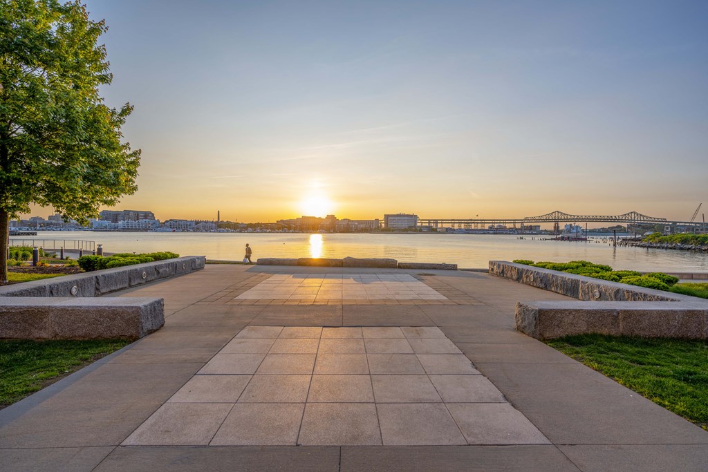 a walkway near the water with the sun setting over the water and a bridge