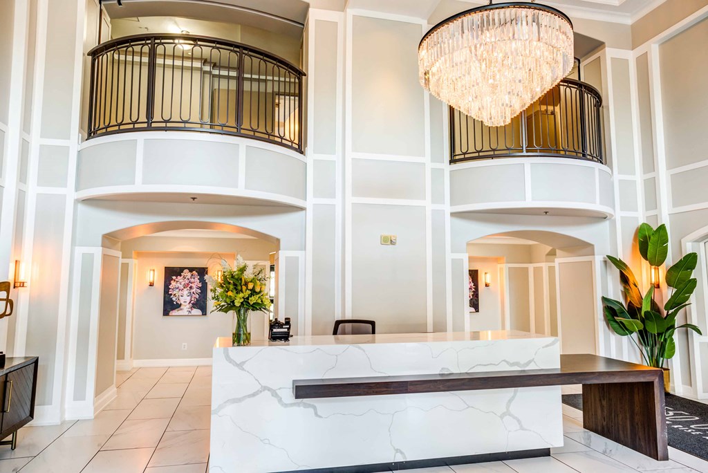 A white reception desk with a marble top is in the center of a spacious lobby with a large chandelier hanging from the ceiling.