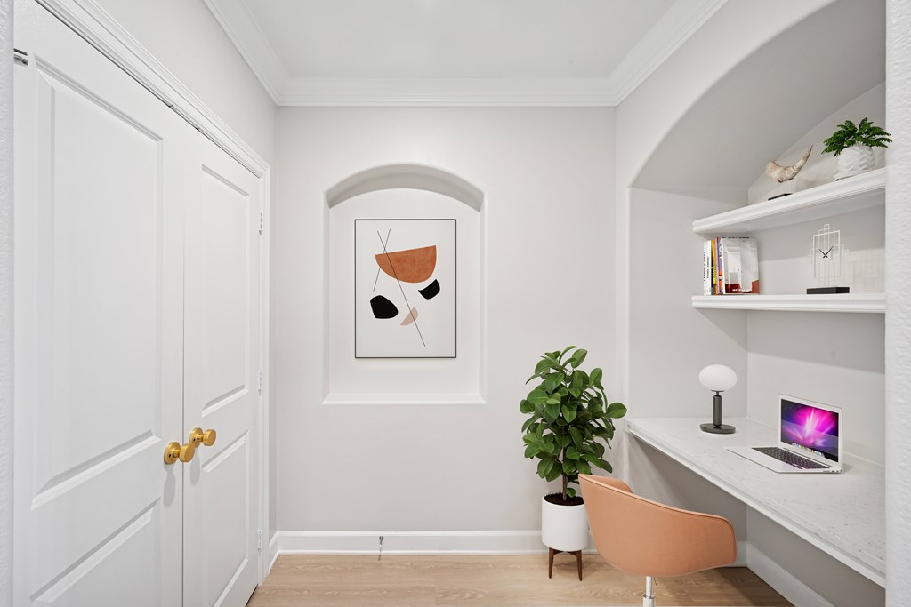 A room with a white door, a painting on the wall, a plant, a chair, a desk with a laptop and a lamp.