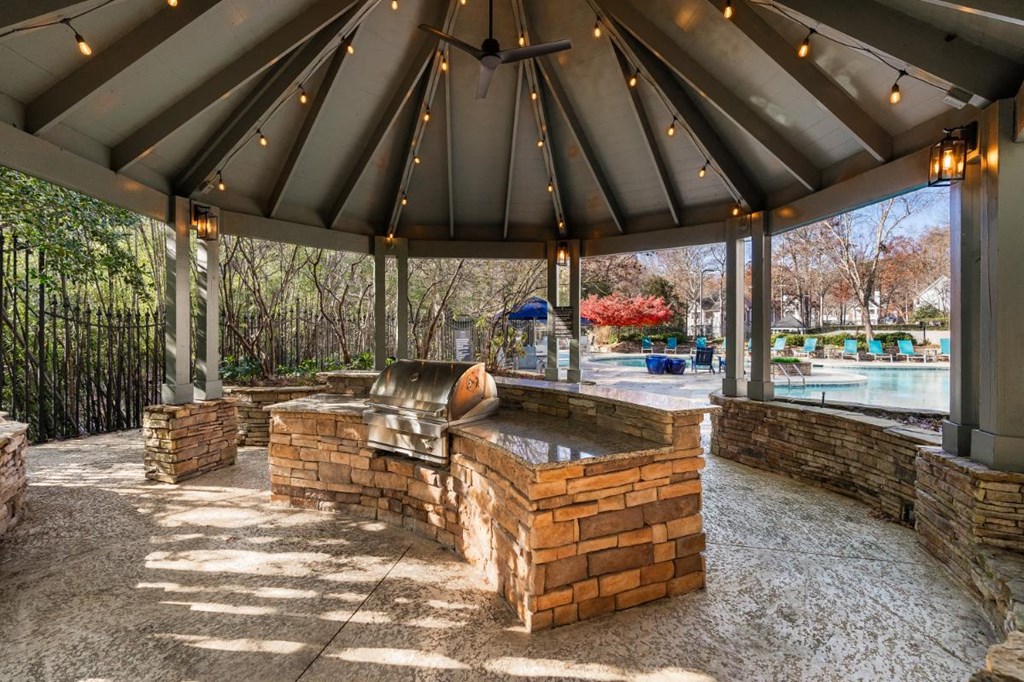 A gazebo with a stone bench and a fireplace is surrounded by a gravel area.