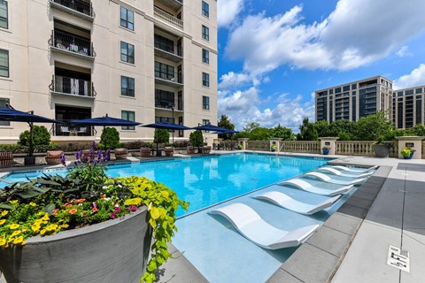 A large swimming pool with a concrete edge and a flower bed in front of a multi-story apartment building.
