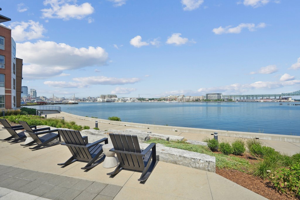 A row of black benches are lined up on a concrete walkway by a body of water.