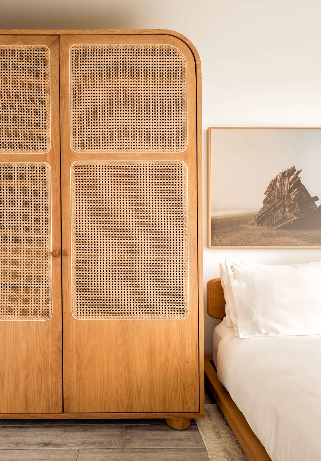 A wooden wardrobe with a perforated design sits next to a bed with a white pillow and a painting of a building in the background.