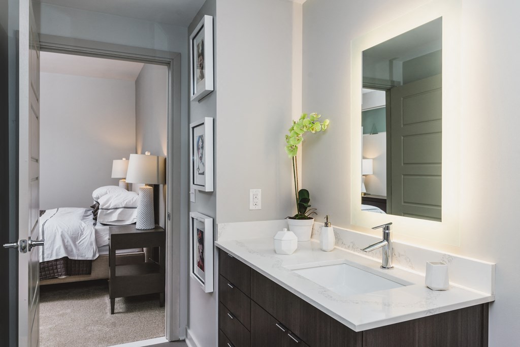 a bathroom with a sink and mirror and a bedroom in the background