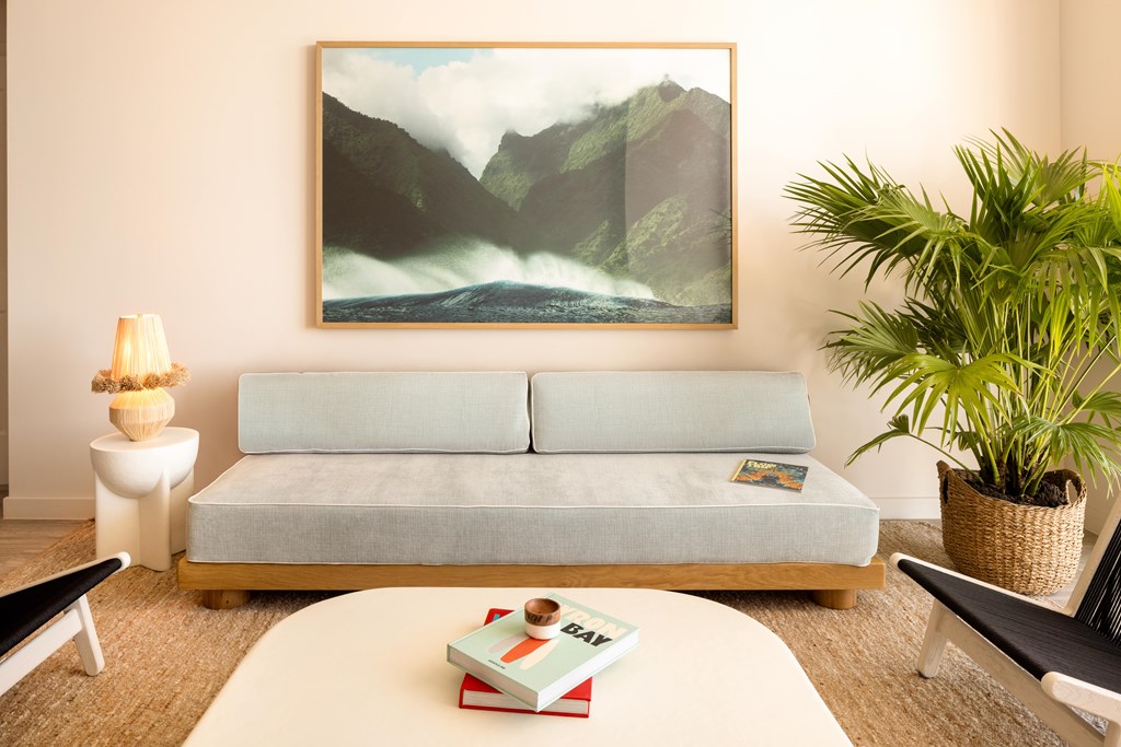 A living room with a grey couch, a painting on the wall, a small table with a lamp and a plant on it, and a white coffee table with a book and a box on it.