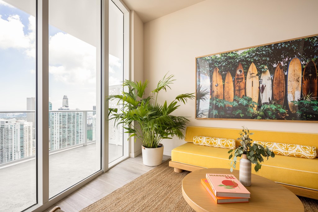 A living room with a yellow couch, a painting on the wall, and a view of the city through the windows.
