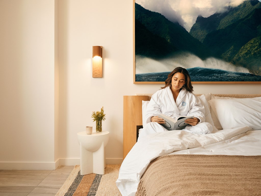 A woman in a white robe is reading a book on a bed with a mountain landscape painting on the wall behind her.