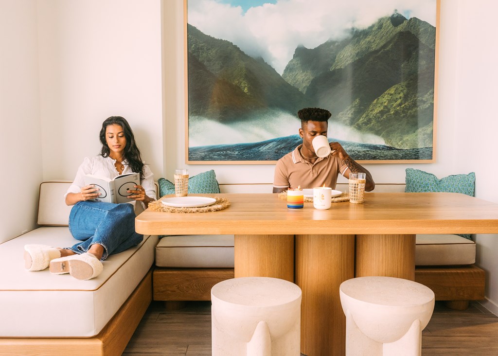 A man and woman are sitting at a table with a large landscape painting behind them.