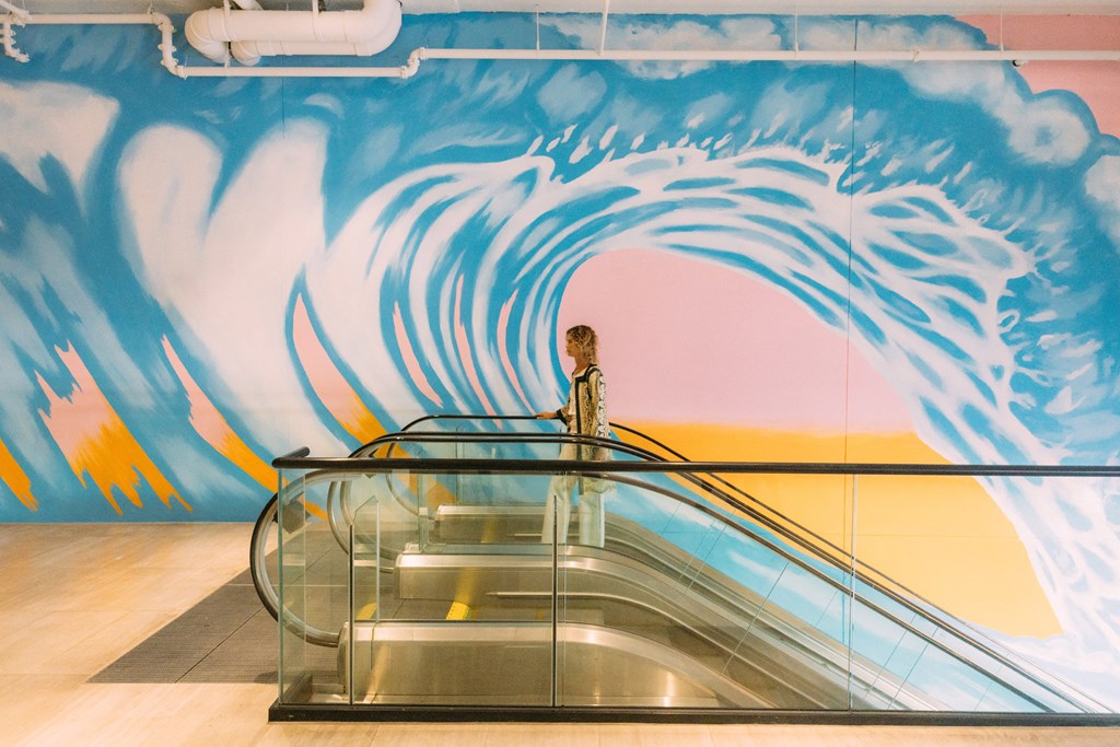 a woman standing at the top of an escalator in front of a wave mural