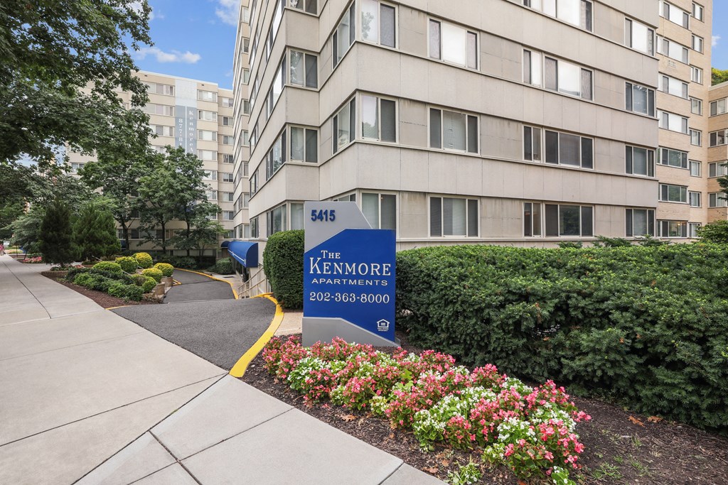 The entrance to The Kenmore Apartments with a blue sign and flowers in the foreground.