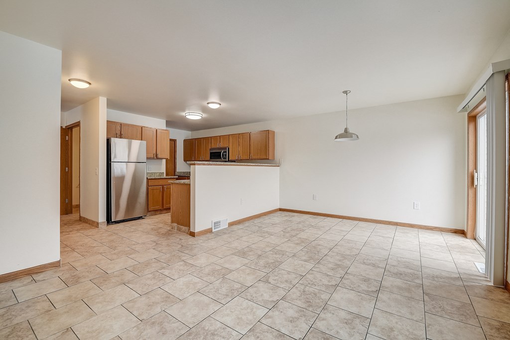 an empty living room with a kitchen with a refrigerator and a sink