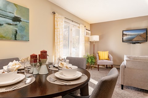 A dining table set with plates and glasses is in the foreground of a living room with a couch and television.