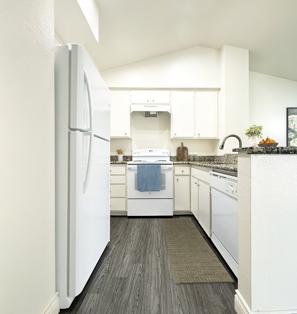 A white refrigerator stands in a kitchen with white cabinets and a white dishwasher at Ridgeline Apartments in San Bernardino