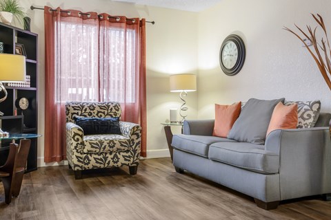 A living room with a grey couch, a patterned chair, and a table with a lamp.