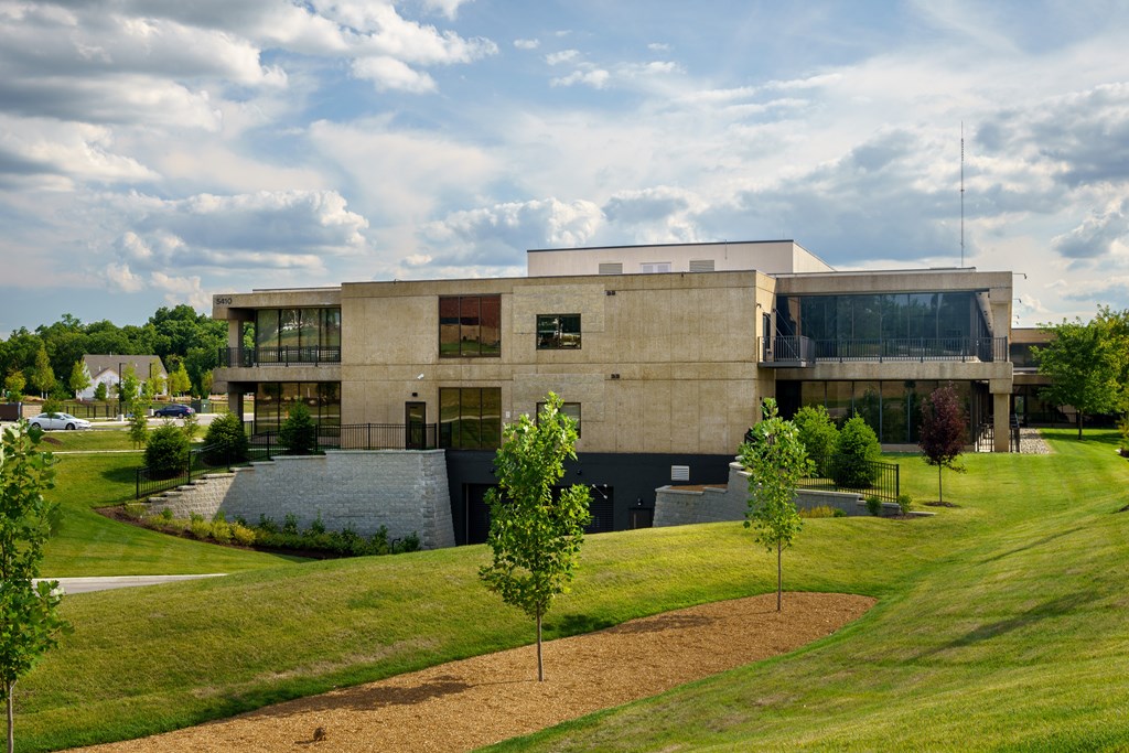 A modern building with a flat roof and large windows is surrounded by a grassy area with trees.