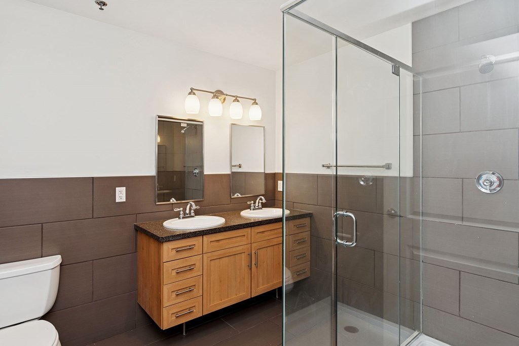 A bathroom with a white toilet, a wooden vanity with two sinks, and a glass shower stall.