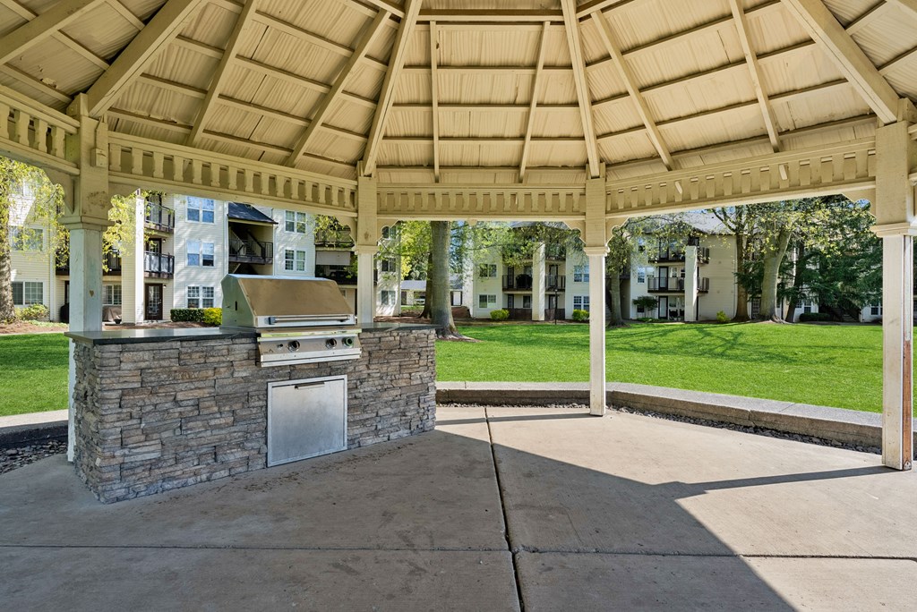A gazebo with a stone pillar and a metal door is surrounded by a concrete floor and a wooden roof.