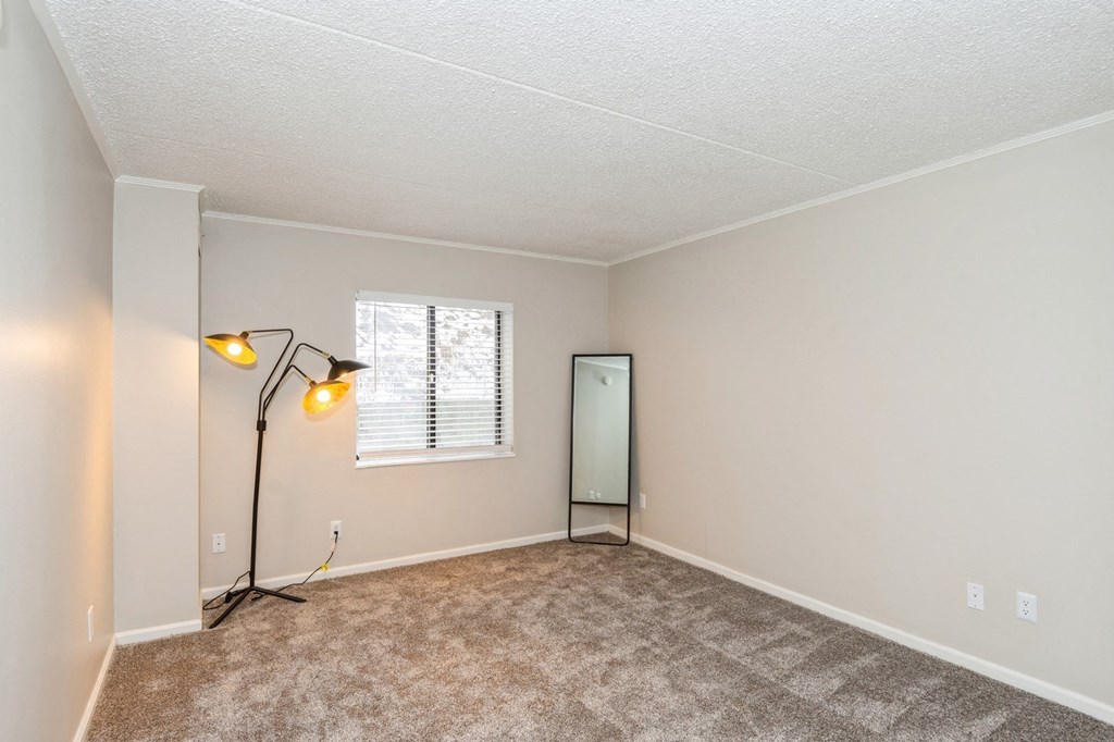A bedroom with a carpeted floor, a window with blinds, a lamp with a yellow shade, and a mirror.