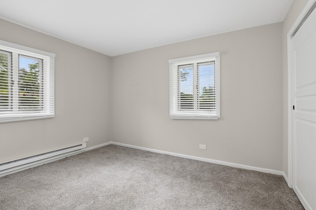 A room with a carpeted floor and two windows with blinds.