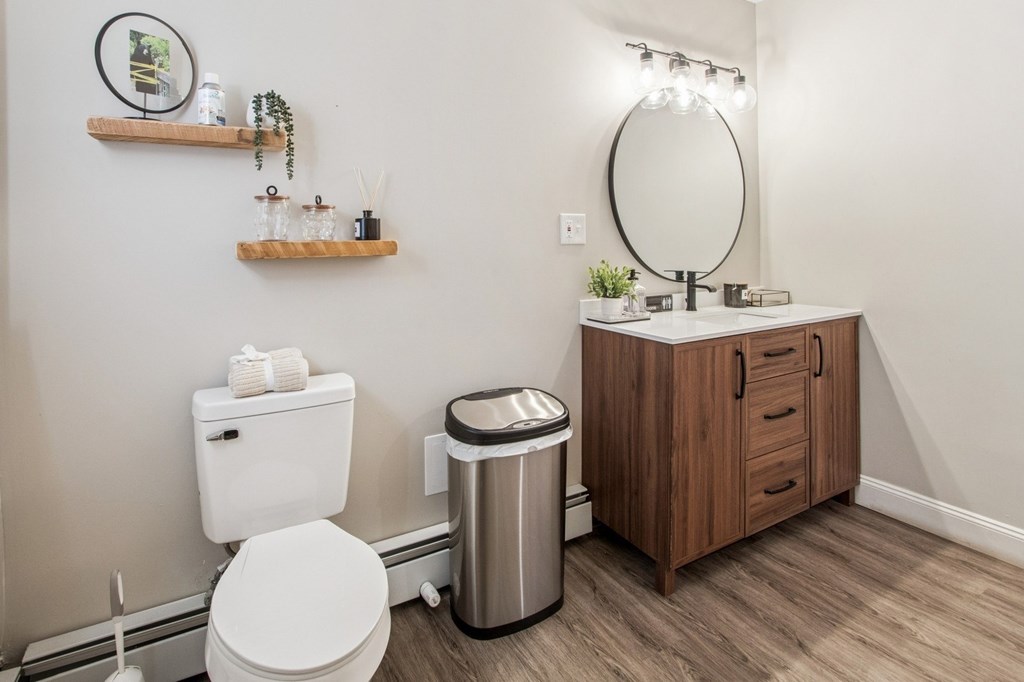 A bathroom with a toilet, a trash can, a mirror, and a wooden cabinet.